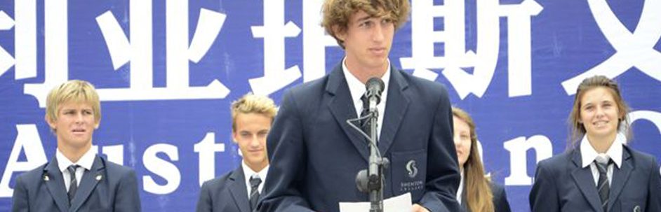 Male senior student talking into a microphone backed by fellow students standing in front of a large blue banner with white Chinese and Latin letters
