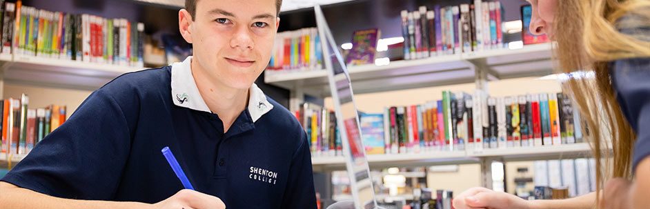 Male student in the library holding a pen and smiling at the camera