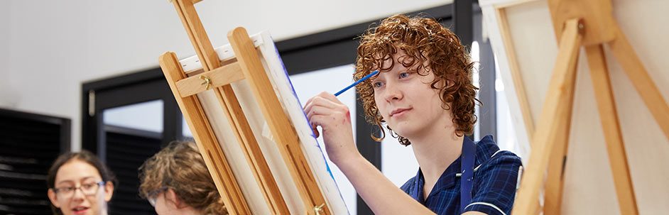 Male student in art class painting on a canvas on an easel with classmates in background