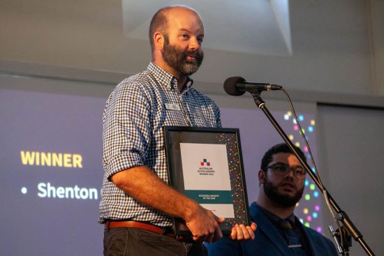 Adam Pengelly award acceptance speech on stage, holding the award frame in front of a microphone