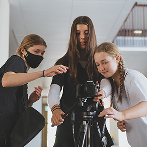 Three students stand gathered around a camera on a stand as they work on a filming project.