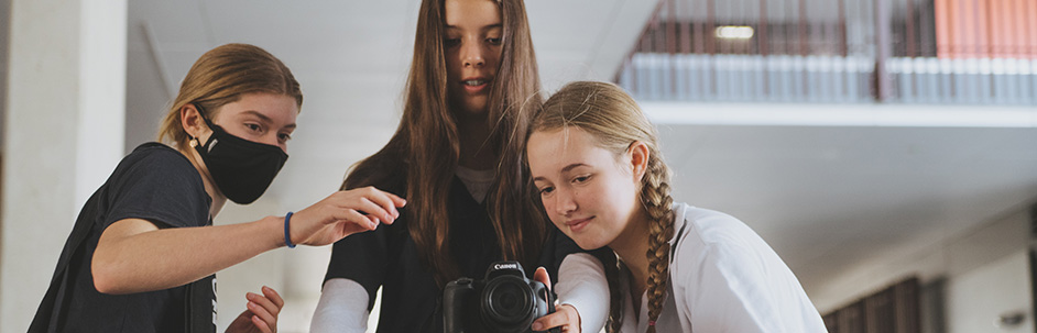 Three students stand gathered around a camera on a stand as they work on a filming project.