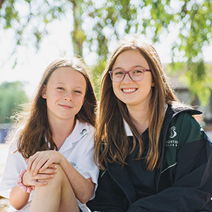 Two students sitting outside on a sunny day smiling at the camera.