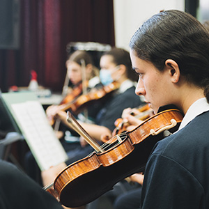 In the foreground is a student playing the violin, with more students playing instruments visible in the background.
